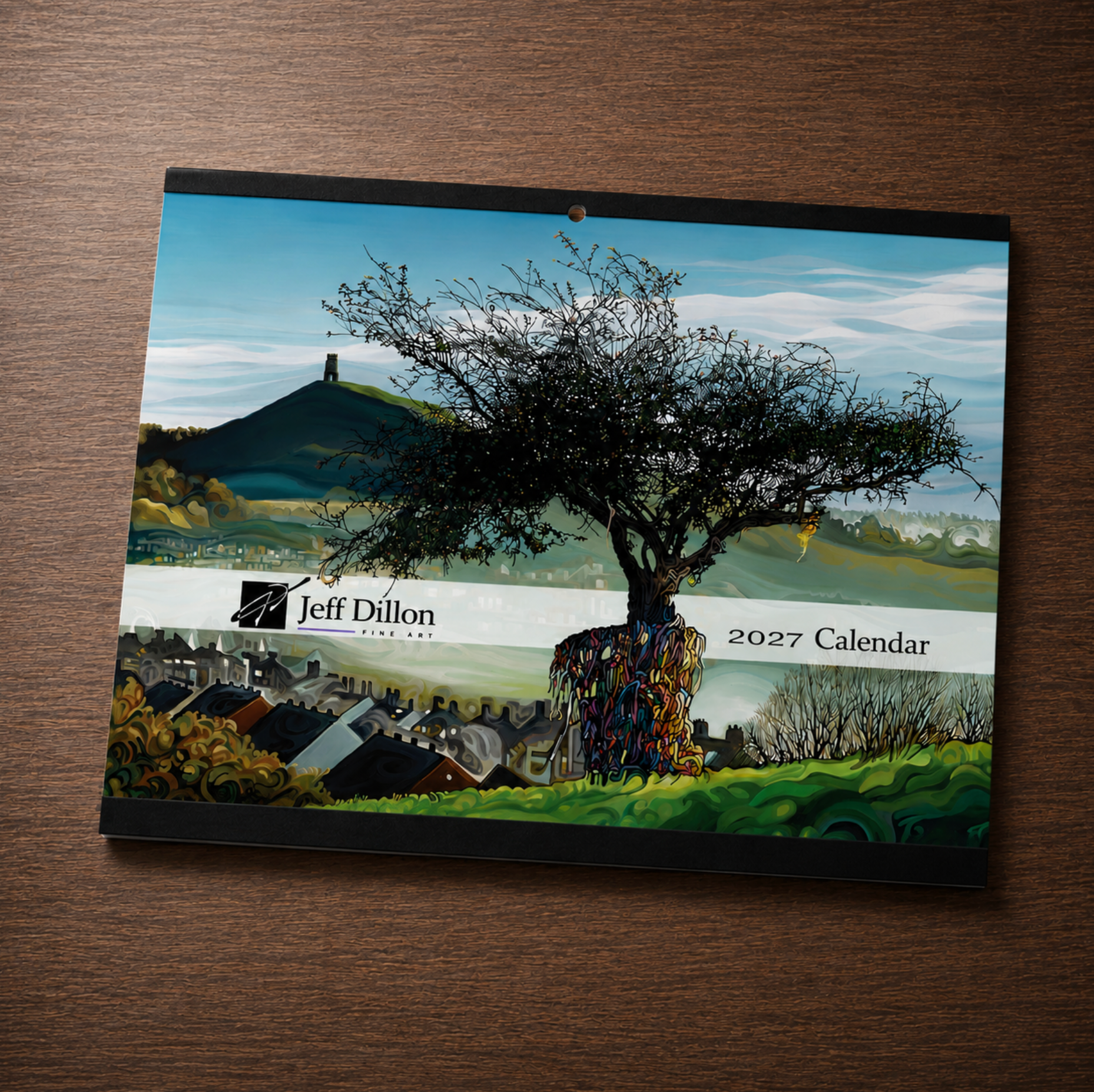A Jeff Dillon 2027 calendar cover displayed flat on a dark wood surface, featuring a landscape painting with a large tree in the foreground, hills, and a village.