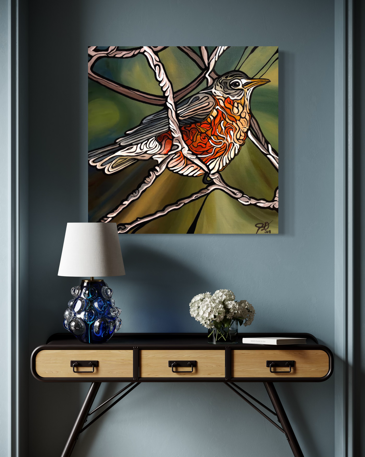 A square canvas art print Jeff Dillon FIne Art depicting a stylized bird with orange, white, and gray feathers on a green and brown background, displayed on a dark gray wall above a console table with a lamp and flowers.