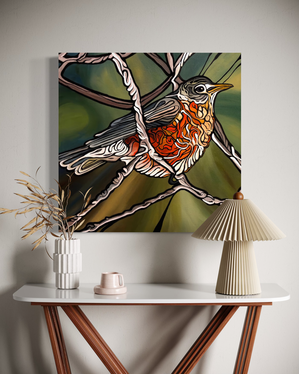 A square wall art print Jeff Dillon FIne Art showing a stylized bird perched on branches, displayed above a console table with a pleated lamp, vase, and small decor object in a neutral interior setting.
