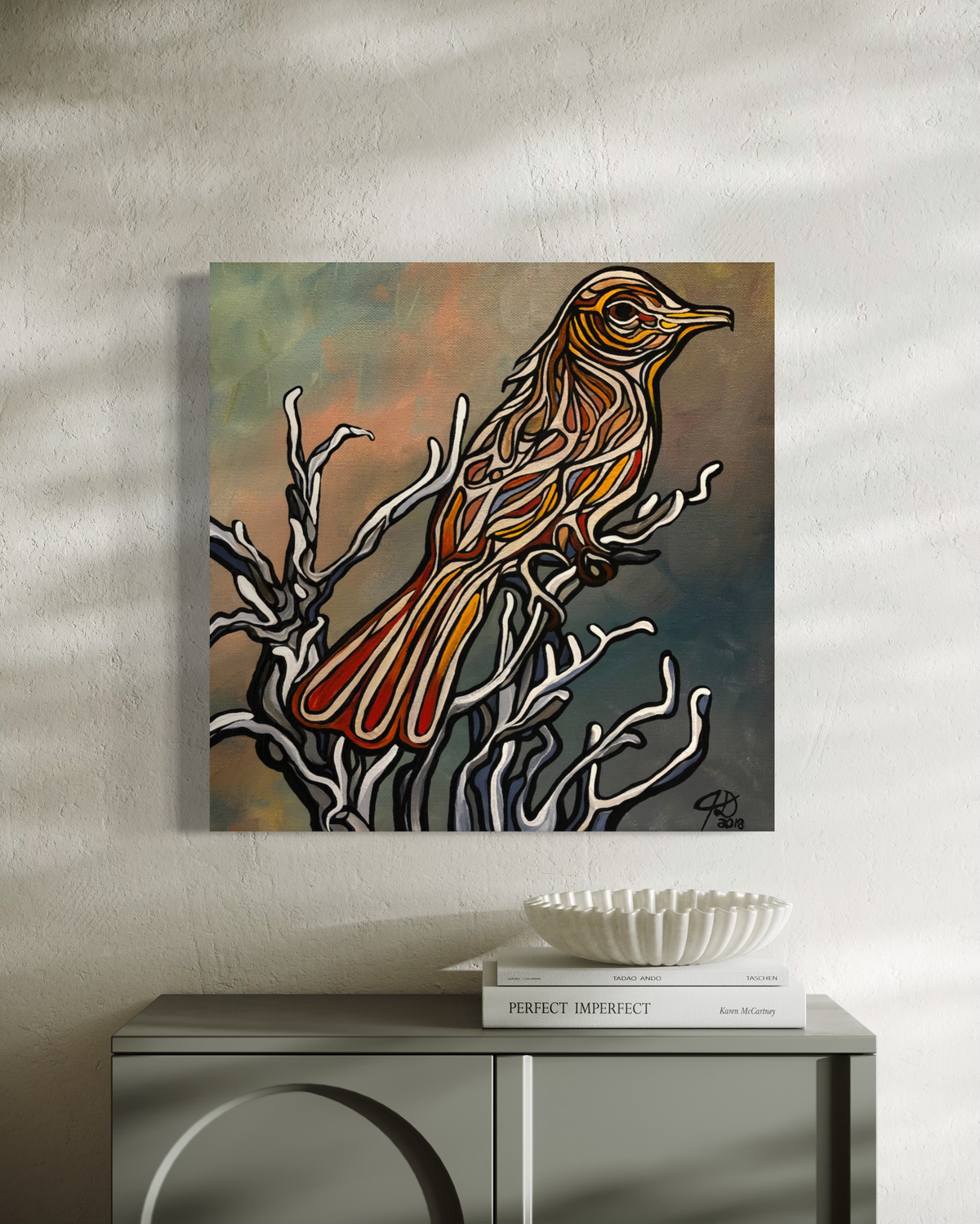 A square canvas art print Jeff Dillon FIne Art featuring a stylized bird perched on branches, displayed on a gray console table against a textured beige wall with soft window shadows.